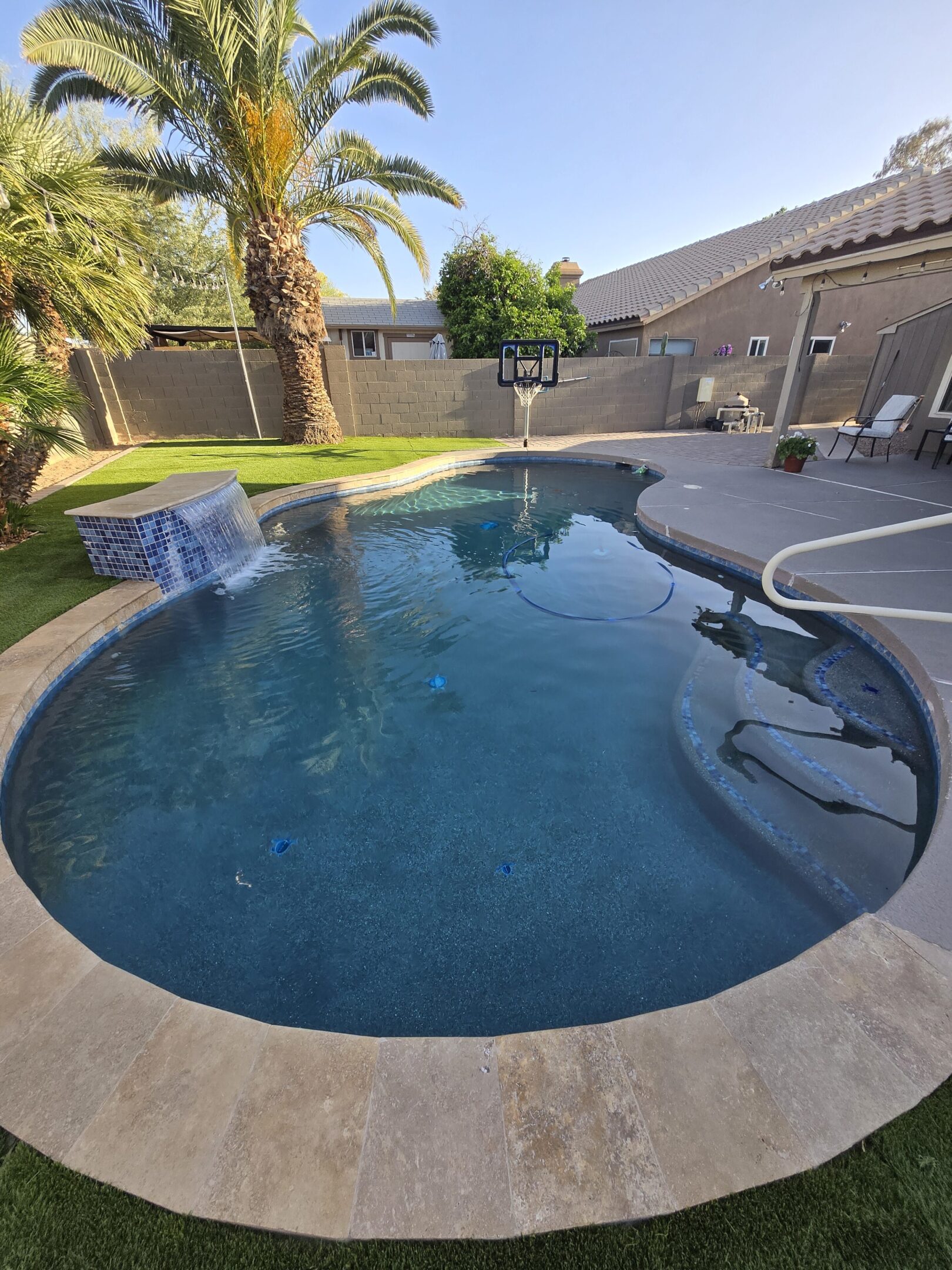 Backyard pool with waterfall and basketball hoop under a clear sky.