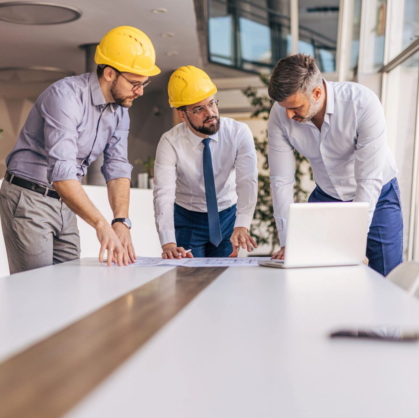 Three professionals in hard hats collaborating over a laptop at a construction site.